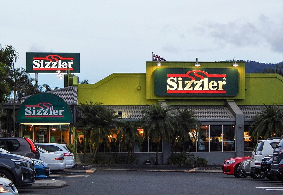 A Sizzler restaurant exterior at dusk, with bright Sizzler signs, palm trees in front, and several cars parked in the parking lot.