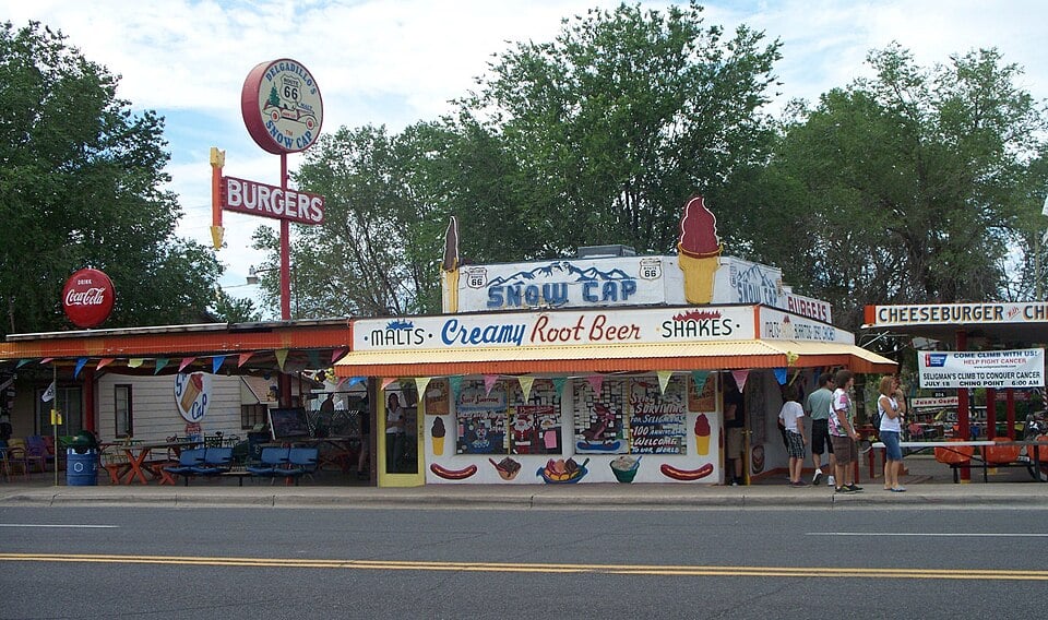 A retro diner with colorful signs for burgers, malts, creamy root beer, and shakes. People stand outside, and there are trees in the background. A large sign and festive banners add to the lively atmosphere.