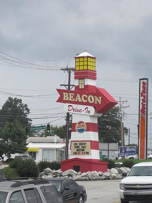 A large lighthouse-shaped sign that reads "BEACON Drive-In" stands by a roadside on a cloudy day, with cars, rocks, and trees visible in the background.