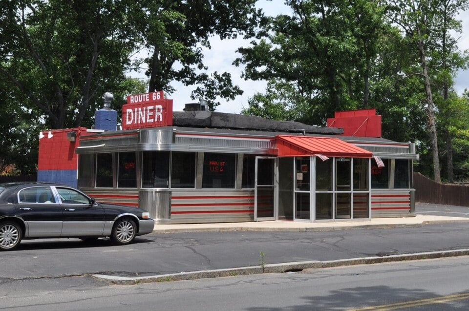 A classic silver diner with red accents and an awning, labeled "Route 66 Diner," sits on a roadside with a car parked nearby and large trees in the background.