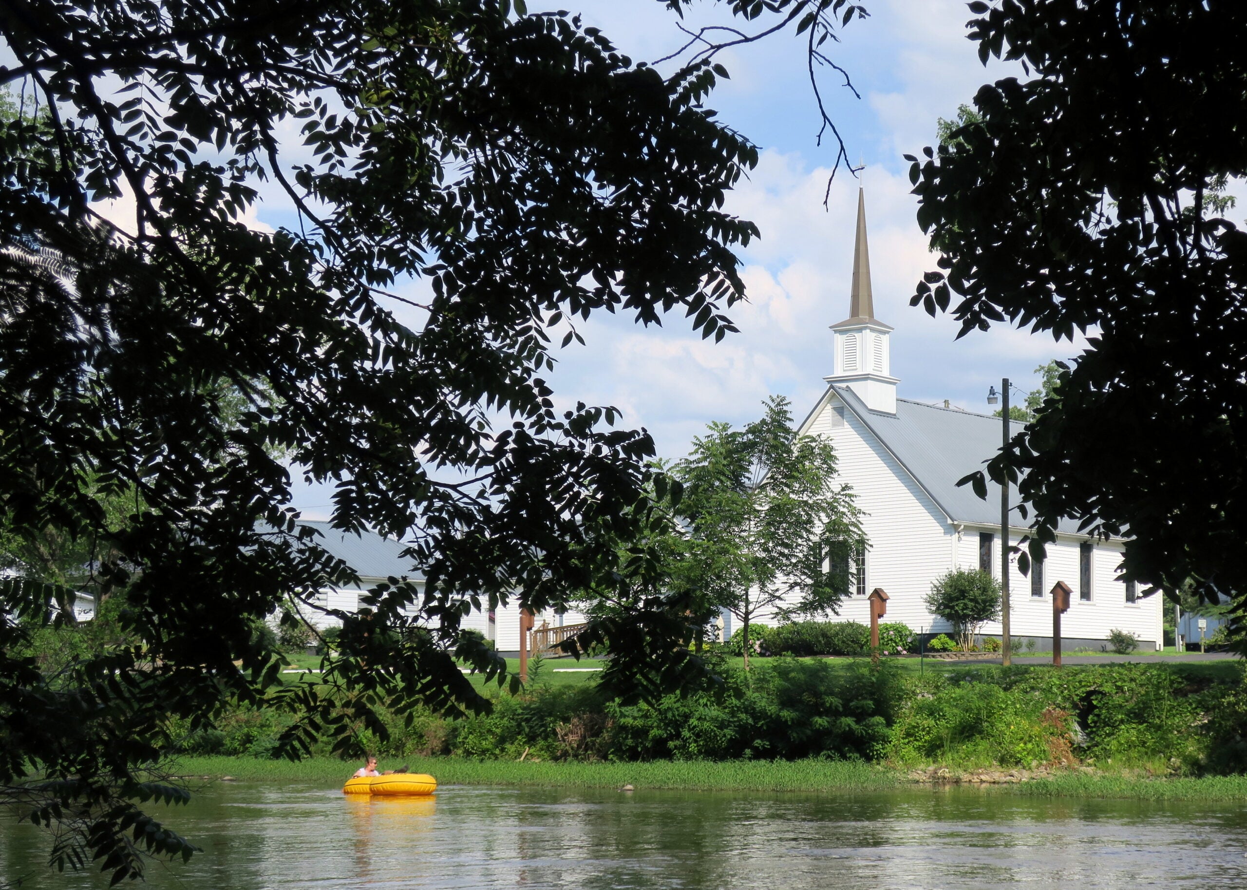 Affordable small towns. A white church with a steeple stands behind trees on a sunny day. In the foreground, a person floats in a yellow inflatable raft on a calm river.