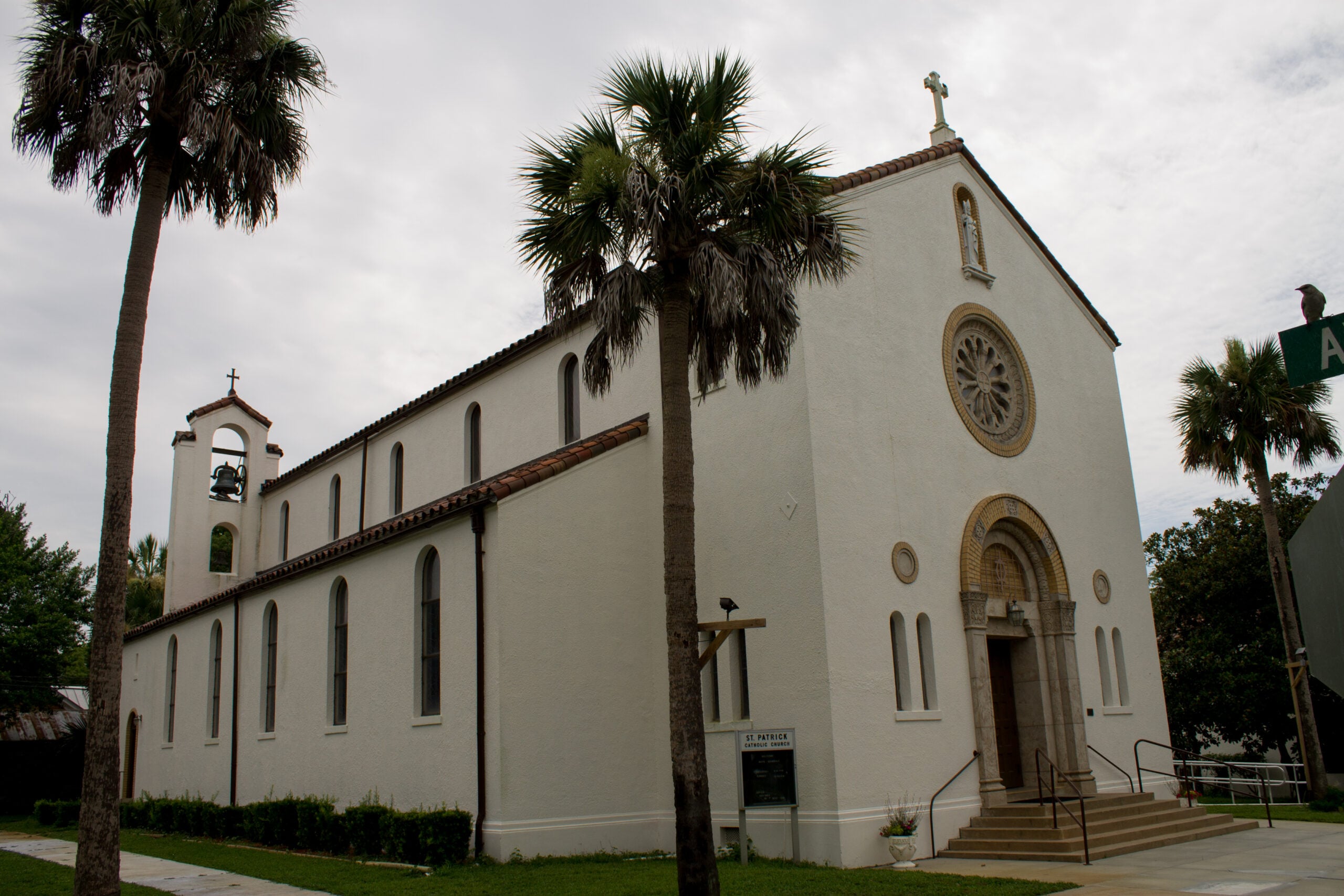 A white stucco church with arched windows, a bell tower, and a circular stained glass window, surrounded by palm trees. Steps lead to the main entrance, and a street sign is visible nearby.