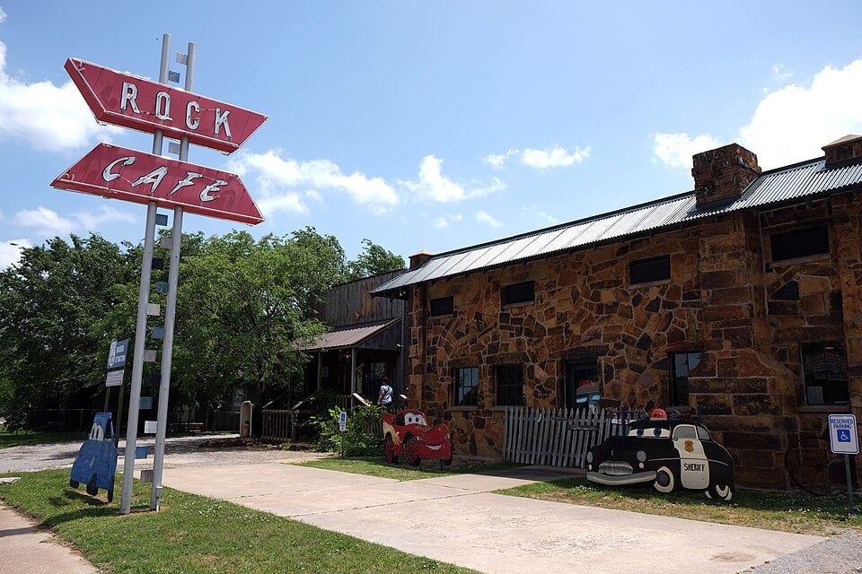 A vintage roadside café with a large red "Rock Café" sign stands next to a stone building. Cartoon car cutouts are displayed outside. The sky is blue with scattered clouds.