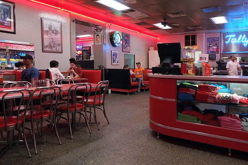 A retro-style diner with red decor, neon lights, and classic metal chairs. Diners sit at tables and booths, while folded t-shirts are displayed on a counter in the foreground. Staff can be seen behind the counter.