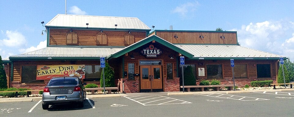 A Texas Roadhouse restaurant with a rustic wooden exterior, metal roof, and front entrance. A car is parked in front and there are signs for parking and an "Early Dine" promotion on the left.