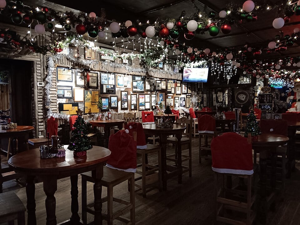 Festively decorated bar with wooden tables and chairs, each chair draped with a Santa hat cover. Christmas lights, ornaments, and garlands hang from the ceiling. Small decorated trees sit on tables; framed photos fill the walls.