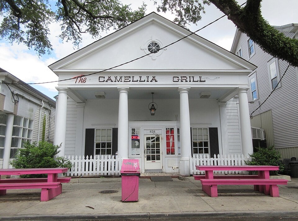 White building with four columns and a triangular pediment reads "The Camellia Grill" above the entrance. Two bright pink benches flank a matching trash bin in front, with a white picket fence and greenery on either side.