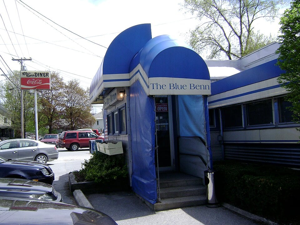Blue awning entrance to The Blue Benn diner, with a visible "Open" sign on the door. Several cars are parked nearby, and a roadside sign with Coca-Cola branding is in the background.