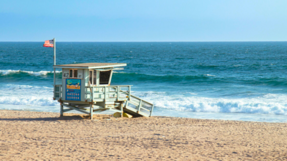 A lifeguard tower with an American flag stands on a sandy beach near the ocean&mdash;one of those cheap family vacation spots where waves crash onto the shore under a clear blue sky, and the calm beach is mostly empty.