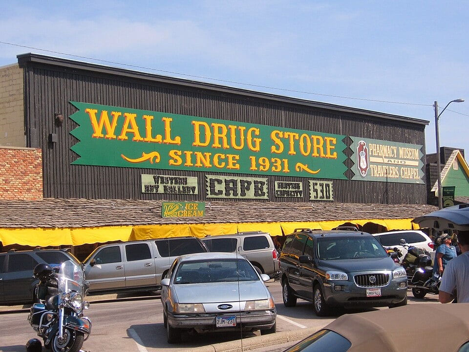 A large sign on a wooden building reads "Wall Drug Store Since 1931" above parked cars and motorcycles. The storefront has yellow awnings, and additional signs advertise ice cream, a pharmacy museum, and a travelers chapel.