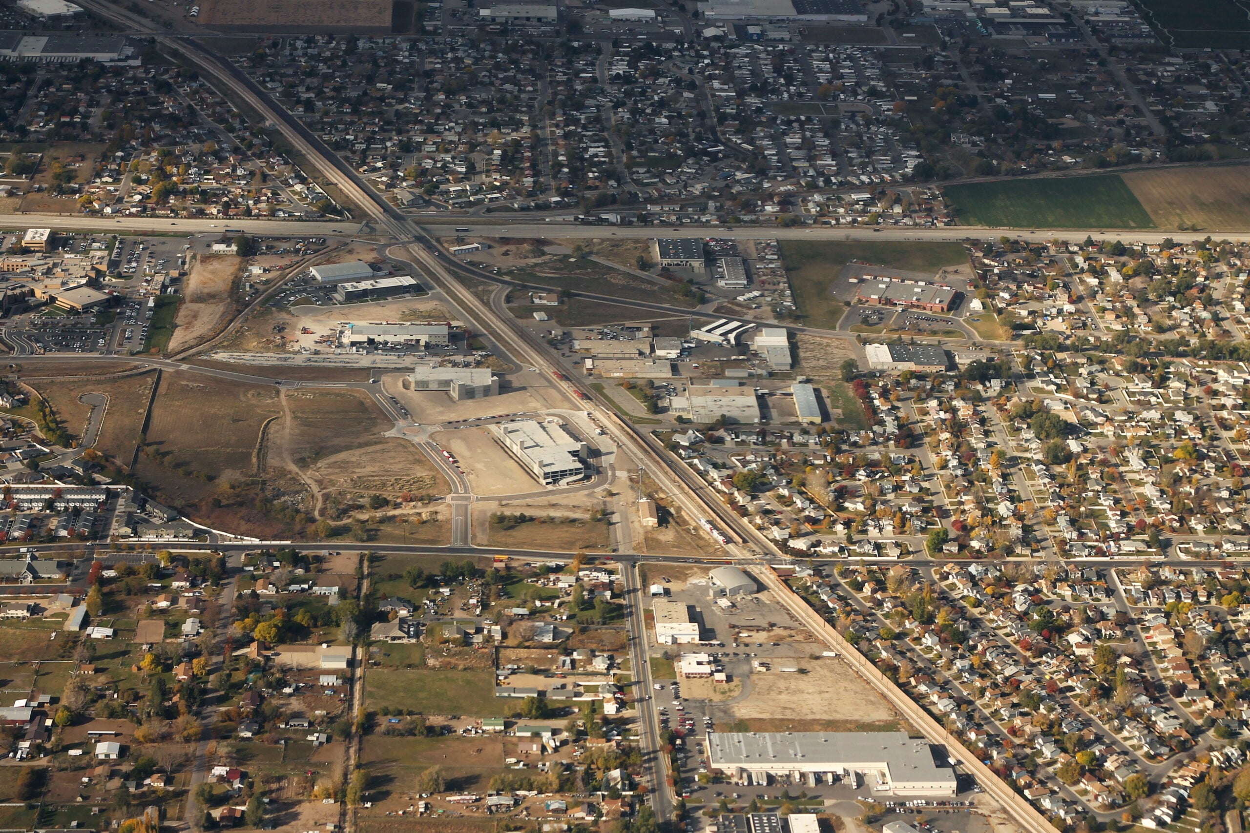 Aerial view of a suburban area with a highway intersection, commercial buildings, parking lots, residential houses, and patches of green spaces. The scene shows a mix of urban and open areas.