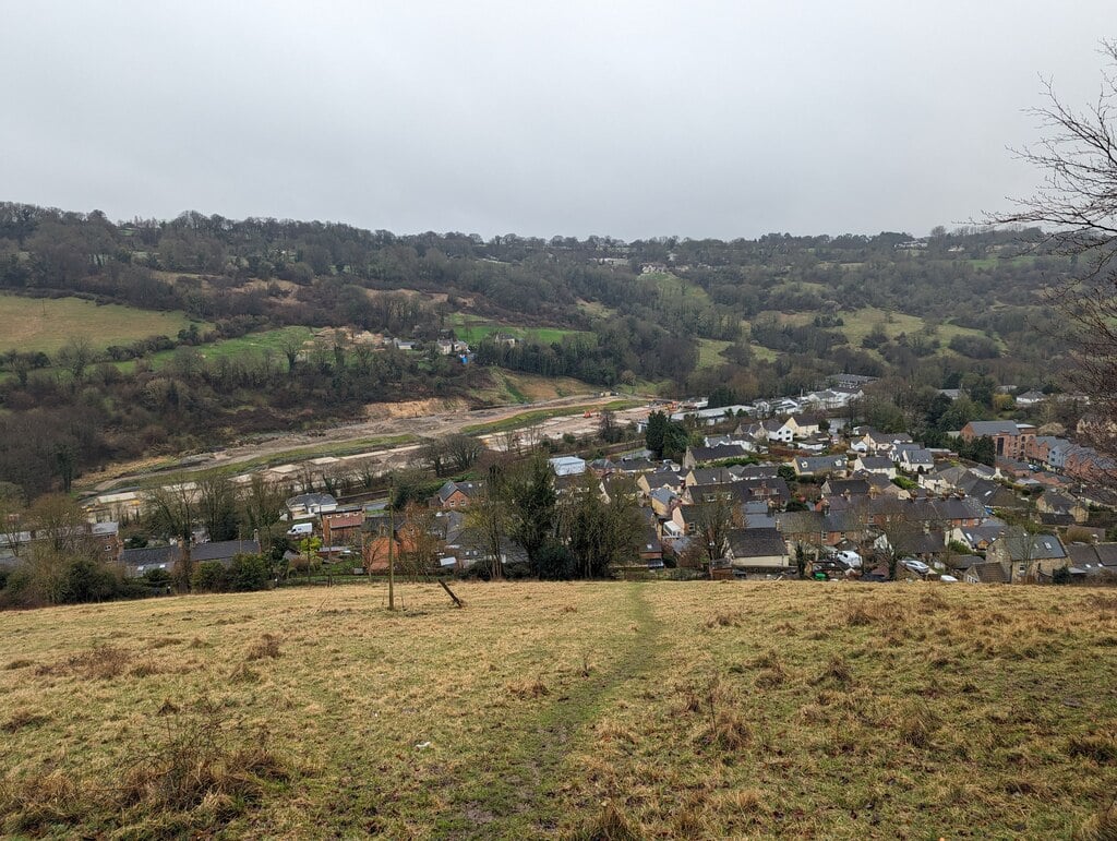 A grassy hill overlooks a small town with houses and buildings, surrounded by green and brown wooded hills under a cloudy, overcast sky. A dirt path leads down the hill toward the town.