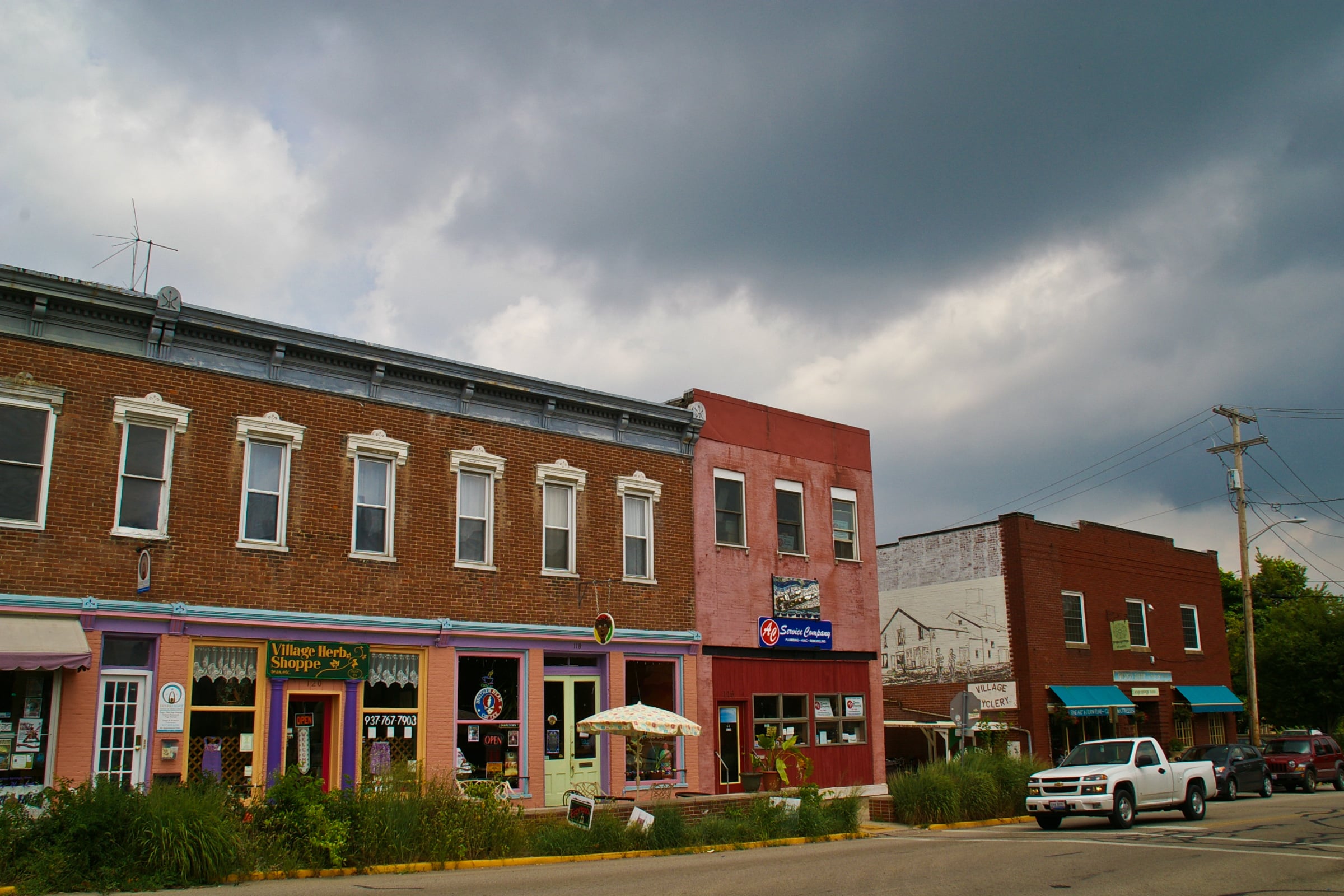A row of colorful small-town storefronts sits under a cloudy, dark sky. A white pickup truck is parked on the street, and some greenery lines the sidewalk. One building has a mural, and a patio umbrella stands out front.