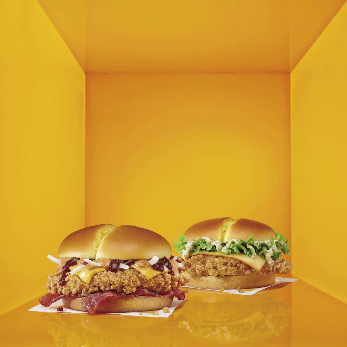 Two crispy chicken sandwiches on square paper trays, set against a bright yellow, box-like background. One Chicken Sandwich features bacon, barbecue sauce, and cheese; the other is topped with lettuce and mayonnaise.