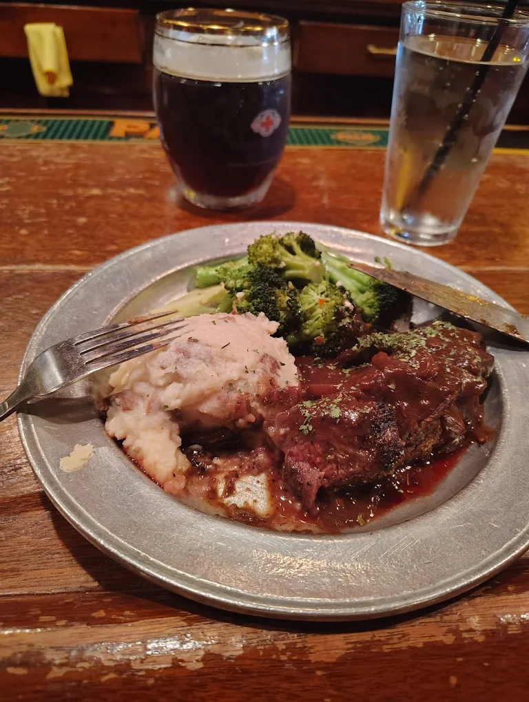 A metal plate with mashed potatoes, steamed broccoli, and beef in gravy on a wooden bar counter. Behind it are a glass of dark beer and a glass of water with a straw.