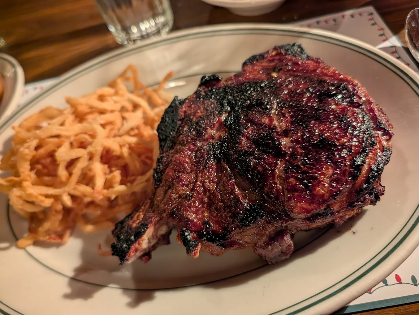 Under-the-radar East Coast steakhouses. A plate with a large, grilled steak that has a charred crust, served next to a pile of thin, crispy fried onions. A glass of water and other dishes are partially visible in the background.
