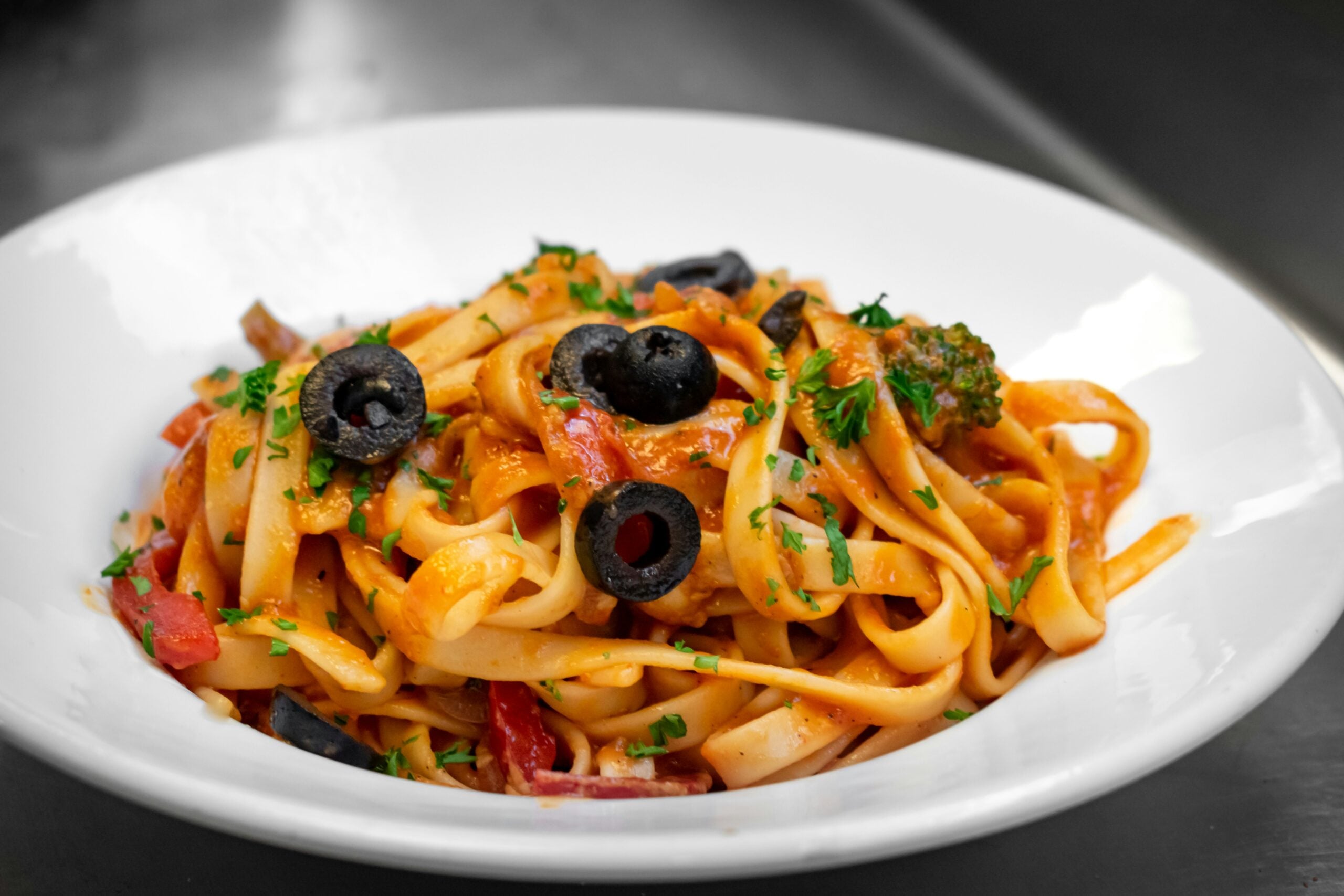 A white bowl filled with fettuccine pasta in a red tomato sauce, garnished with sliced black olives, chopped parsley, and mixed vegetables.