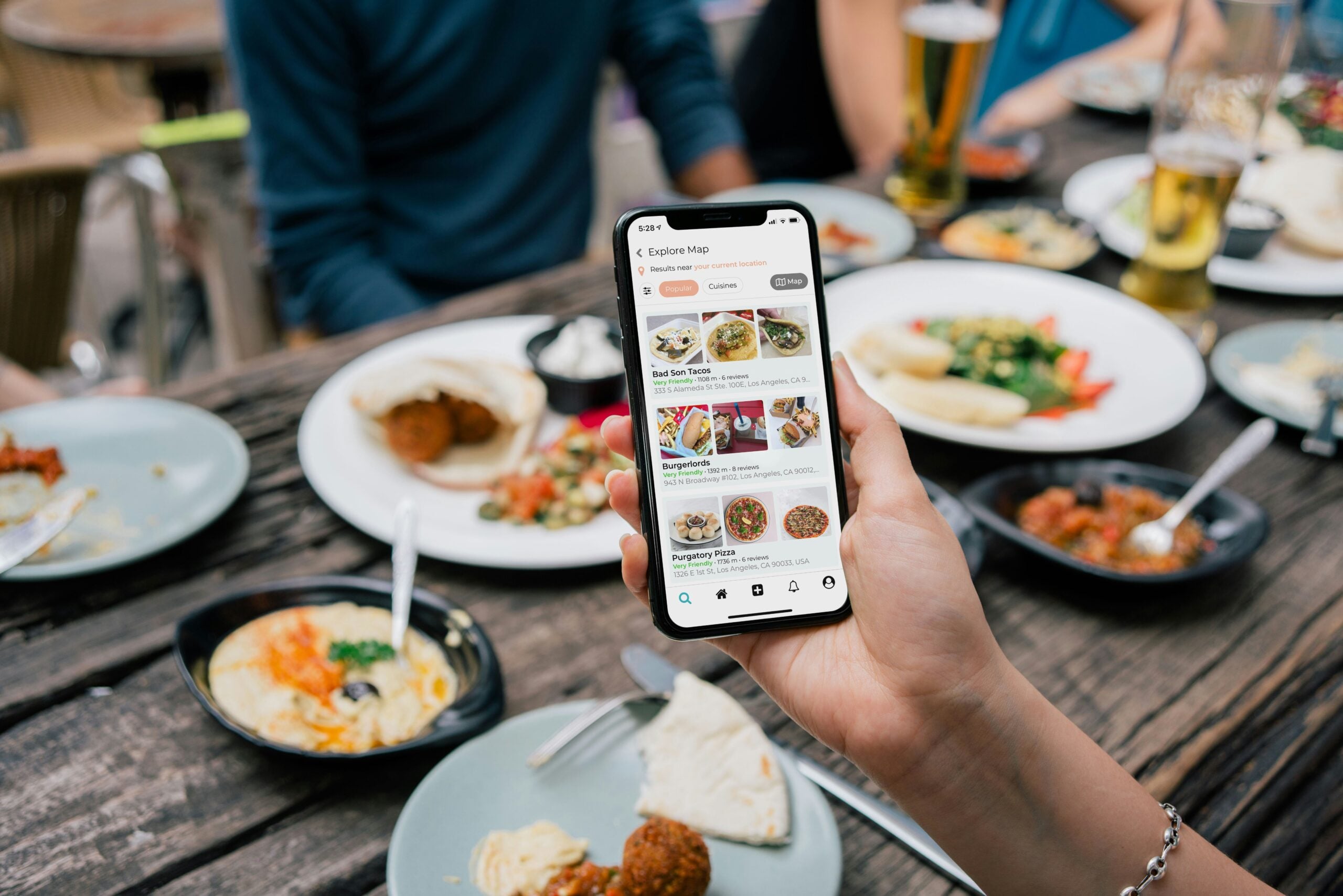 A person holds a smartphone displaying a food delivery app while sitting at a wooden table set with various plates of food, drinks, and partially eaten meals, surrounded by other diners.