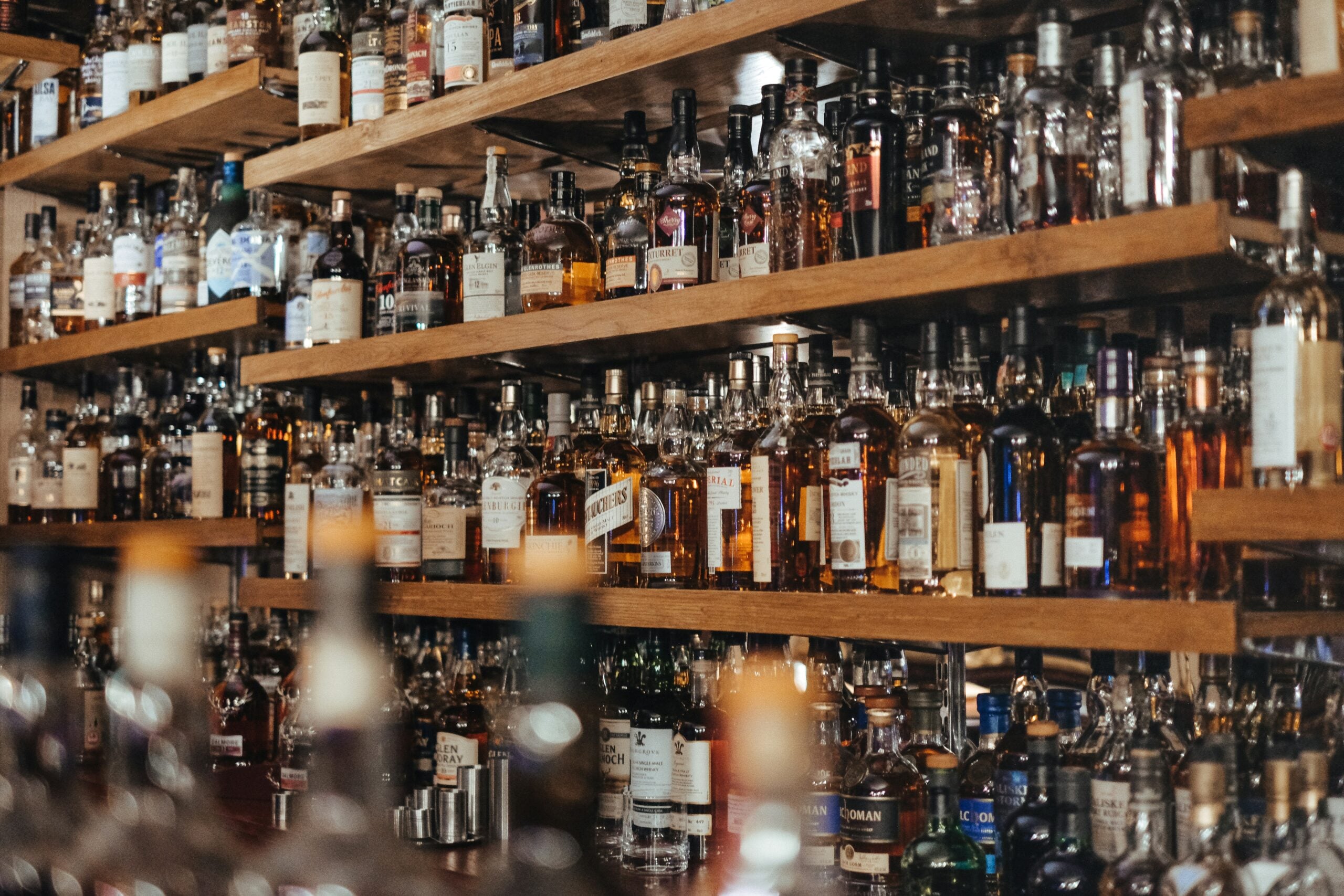 Shelves filled with numerous bottles of liquor and spirits in a bar or liquor store, with various shapes, sizes, and labels on display. The scene is busy and colorful with a wide selection of drinks.