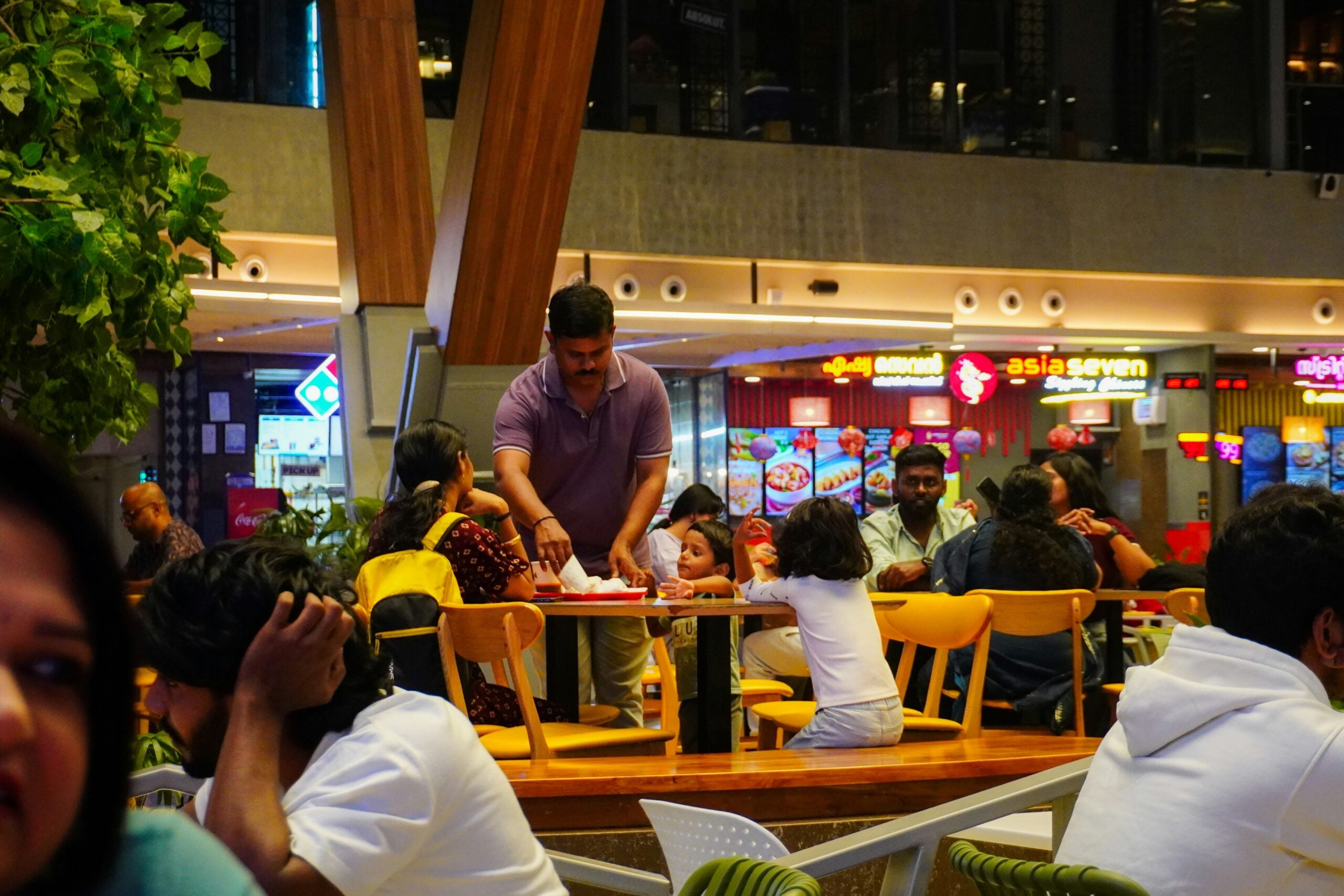 A busy food court with several people sitting at tables. A man in a purple shirt serves food to a woman and two children at a table. Bright lights and food counters are visible in the background.