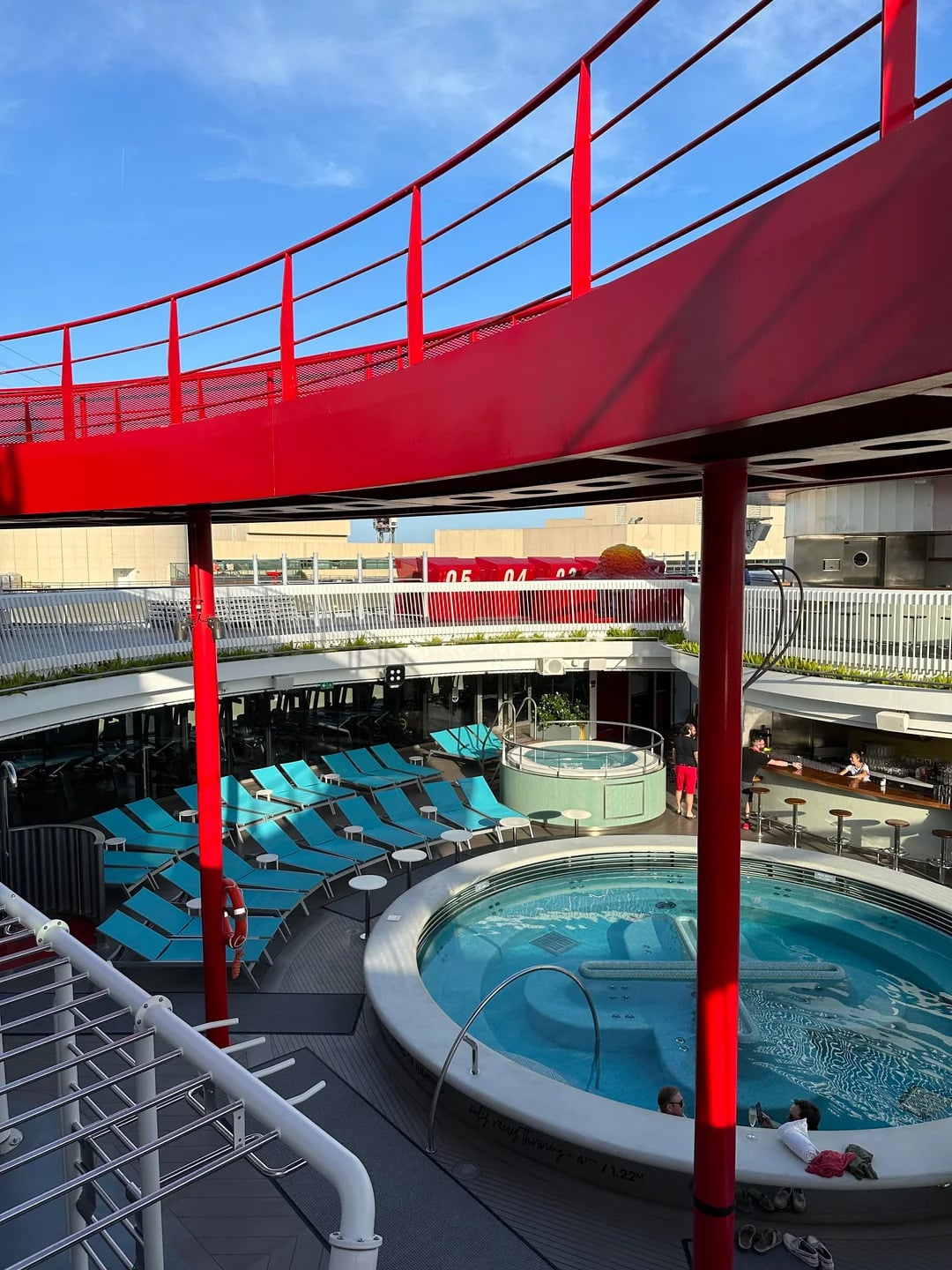 A circular hot tub and light blue lounge chairs surround a pool deck on a modern cruise ship, framed by bright red railings and set against a clear blue sky—experience the Best Cruise Line 2026 in style.
