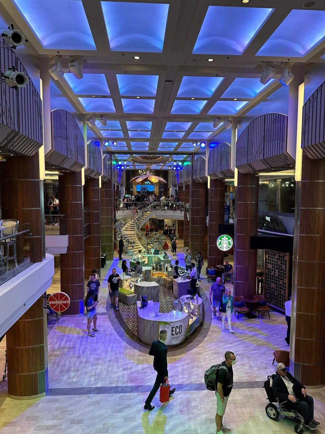 A spacious, brightly lit indoor promenade on the Best Cruise Line 2026 features cafes, shops, and people relaxing under blue ceiling lights, with a Starbucks kiosk and a modern, vibrant atmosphere.