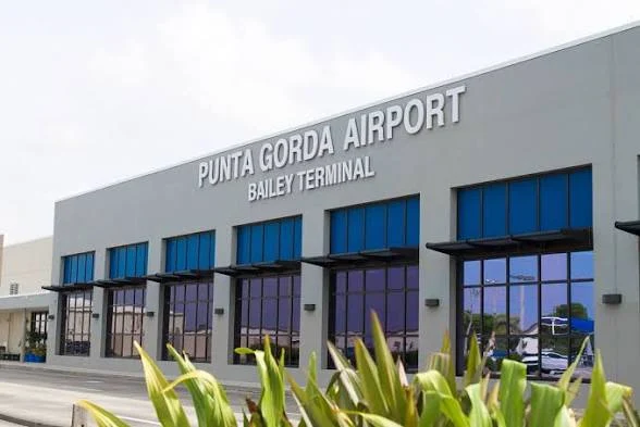 Exterior view of Punta Gorda Airport’s Bailey Terminal, featuring large blue-tinted windows and clear signage on the building. Some greenery is visible in the foreground.