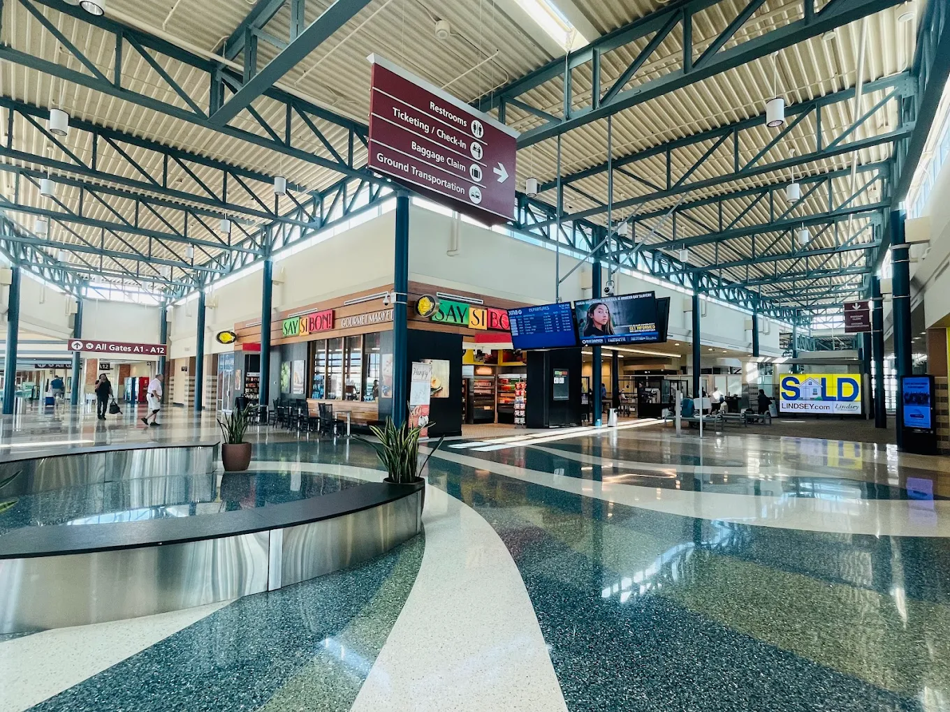 Spacious airport terminal with high ceilings, decorative floor patterns, shops, and restaurants. Overhead signs direct travelers, and digital flight information screens are visible. Natural light fills the area.