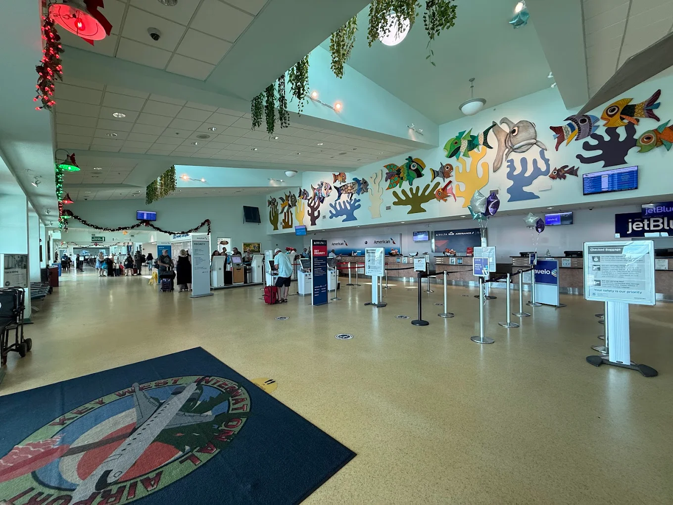 A brightly lit airport check-in area with JetBlue counters, colorful sea-themed wall art, people waiting in line, flight info screens, and overhead hanging plants. The floor has a large airport-themed rug.