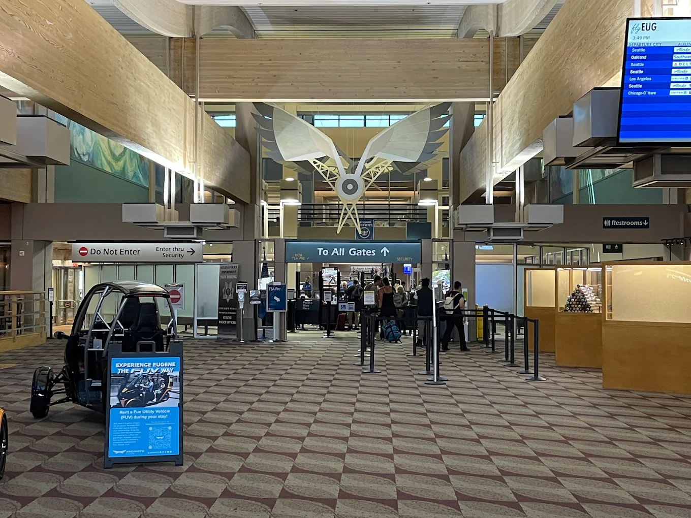 Airport terminal interior with passengers in line at security, large stylized silver bird sculpture above, check-in signs, and information displays visible. Patterned carpet and restroom sign on the right side.