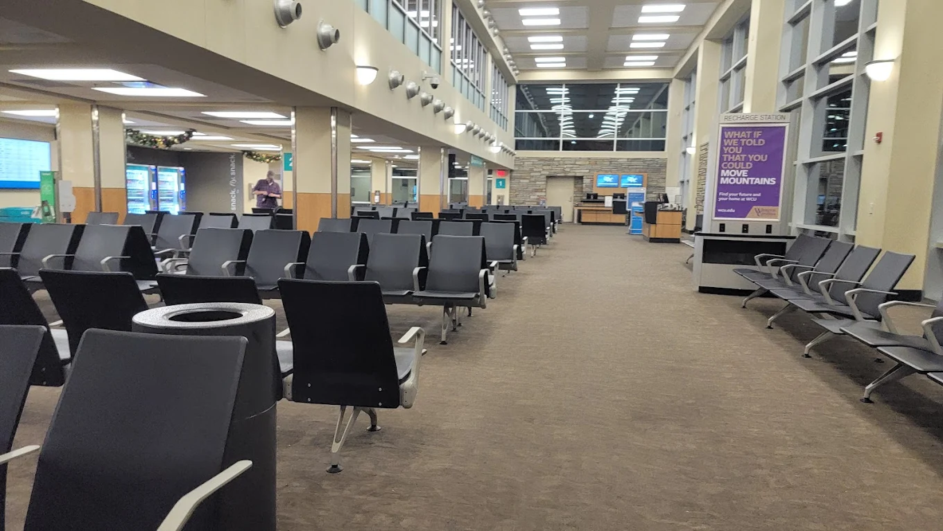 A nearly empty airport waiting area with rows of black chairs, a trash can, and large windows. A person stands near a counter in the distance, and a purple sign is visible on the right side.
