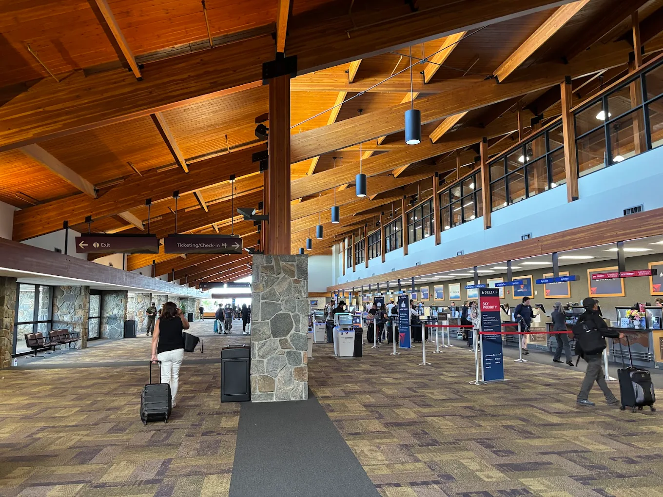 A spacious airport terminal with wooden ceilings, stone pillars, and people walking or waiting in line at check-in counters; luggage and signs are visible throughout the area.