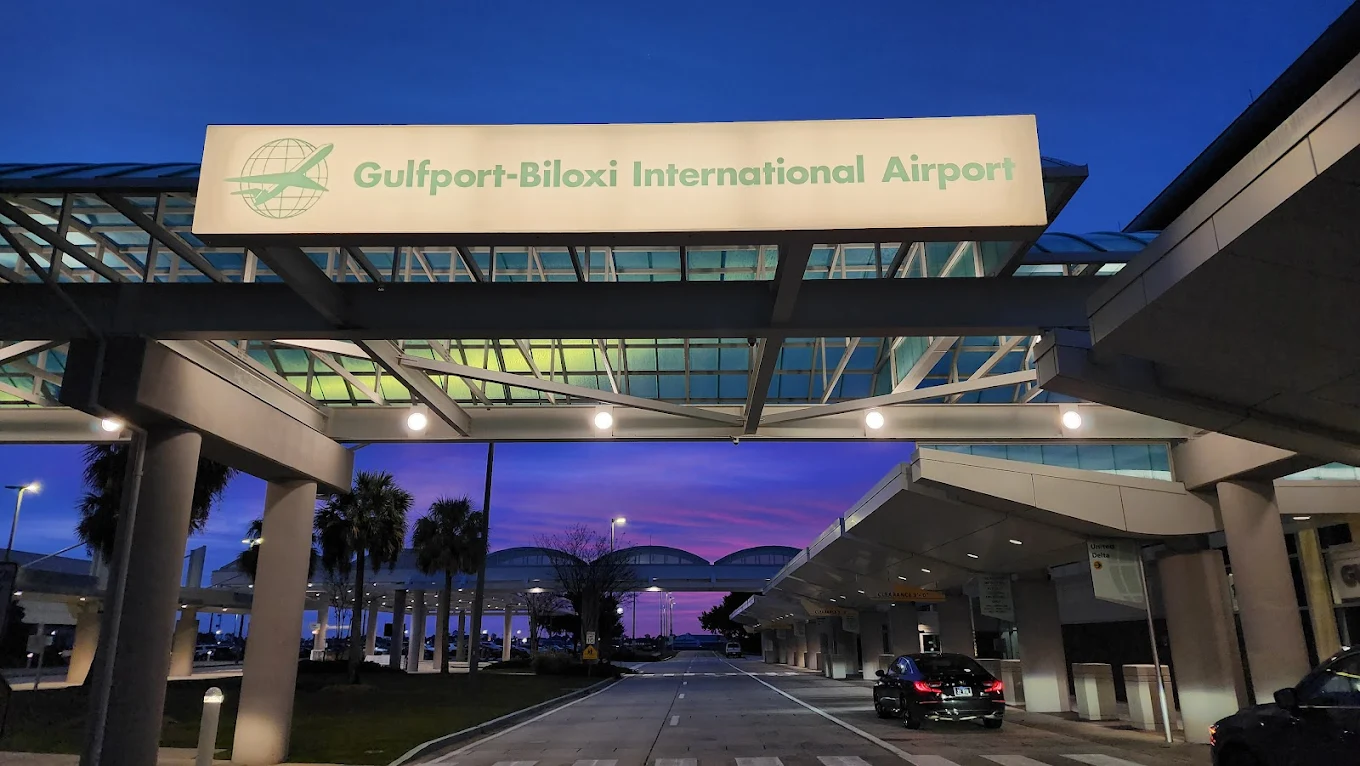 Entrance to Gulfport-Biloxi International Airport at dusk, with a lit sign overhead, modern glass canopy, palm trees, and cars parked at curbside under a deep blue and purple sky.