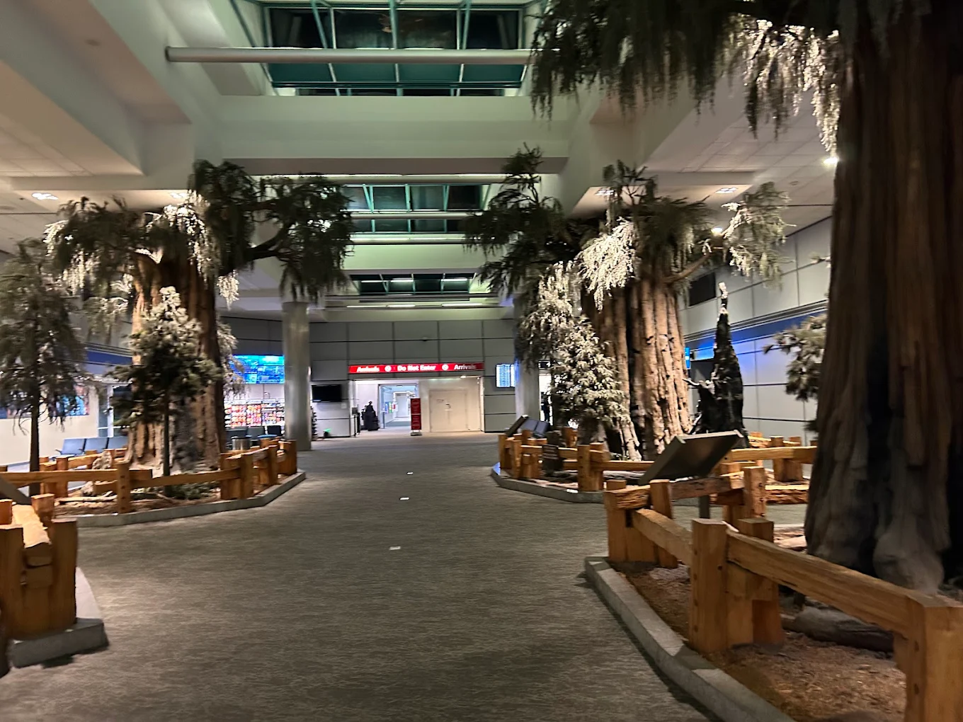 An indoor airport walkway features large artificial trees, rustic wooden fences, and dim lighting, creating a forest-like atmosphere. Signs and seating areas are visible in the background.
