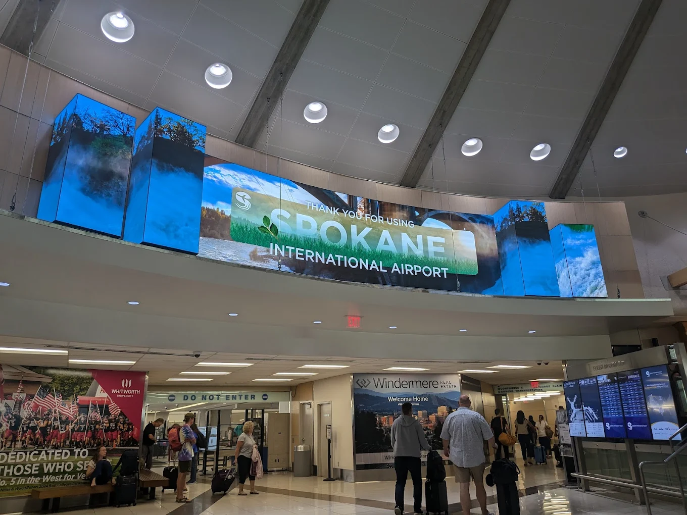A large digital sign reads "Thank you for using Spokane International Airport" above a busy airport terminal with travelers walking and checking monitors.