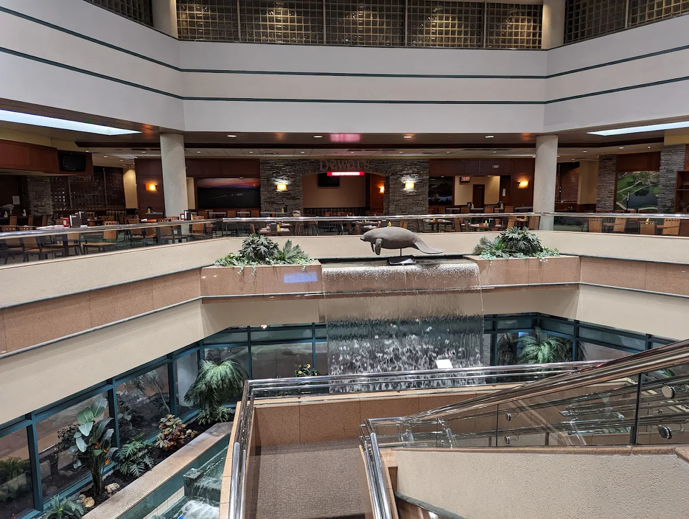 Interior of a mall or atrium with a cascading waterfall, glass railings, an upper dining area with tables and chairs, and a sculpture of a seal above the waterfall.