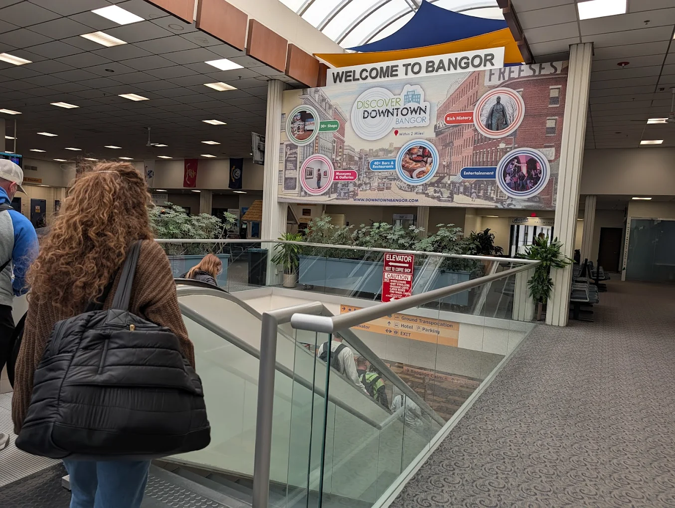 A woman with curly hair and a black bag walks past a large "Welcome to Bangor" banner inside an airport, featuring images and attractions from downtown Bangor. Other travelers and plants are visible in the terminal. Affordable vacations from regional airports.