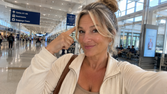 A woman with blonde hair in a bun smiles and points to her temple, sharing her favorite airport hacks while taking a selfie in a bright, spacious airport terminal with signs for gates, restrooms, and baggage claim visible overhead.