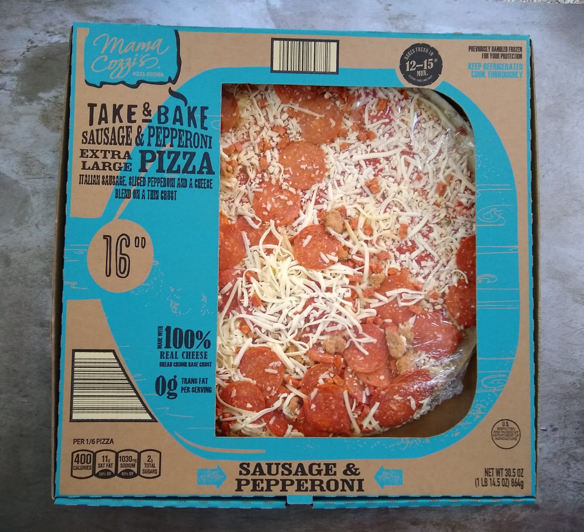 A Mama Cozzi's Take & Bake Sausage & Pepperoni Extra Large Pizza in a cardboard box, topped with shredded cheese, sausage, and pepperoni, shown on a gray countertop.