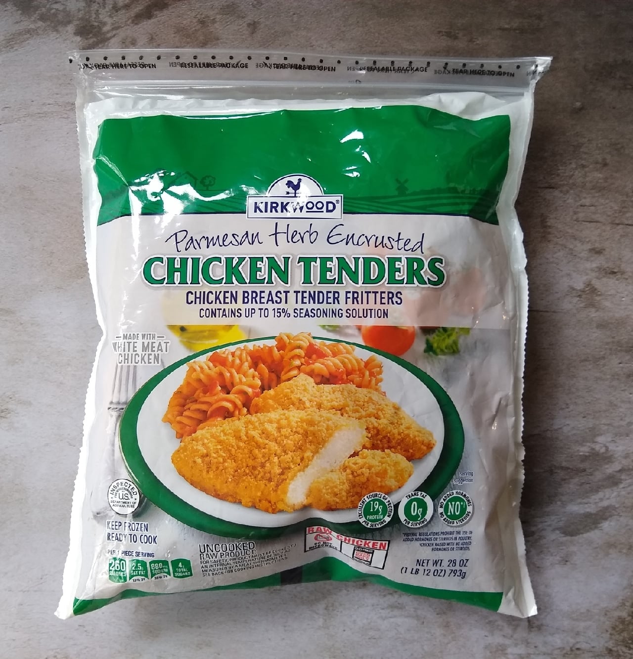 A bag of Kirkwood Parmesan Herb Encrusted Chicken Tenders is placed on a grey surface. The package shows an image of breaded chicken tenders with pasta and vegetables on a plate.