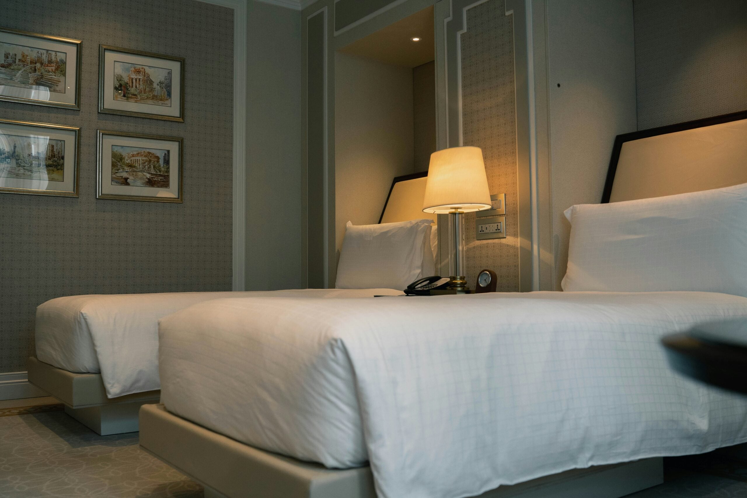Two neatly made twin beds with white linens in a hotel room. A lit bedside lamp, phone, and clock are on the nightstand between the beds. Four framed pictures hang on the patterned wall to the left.