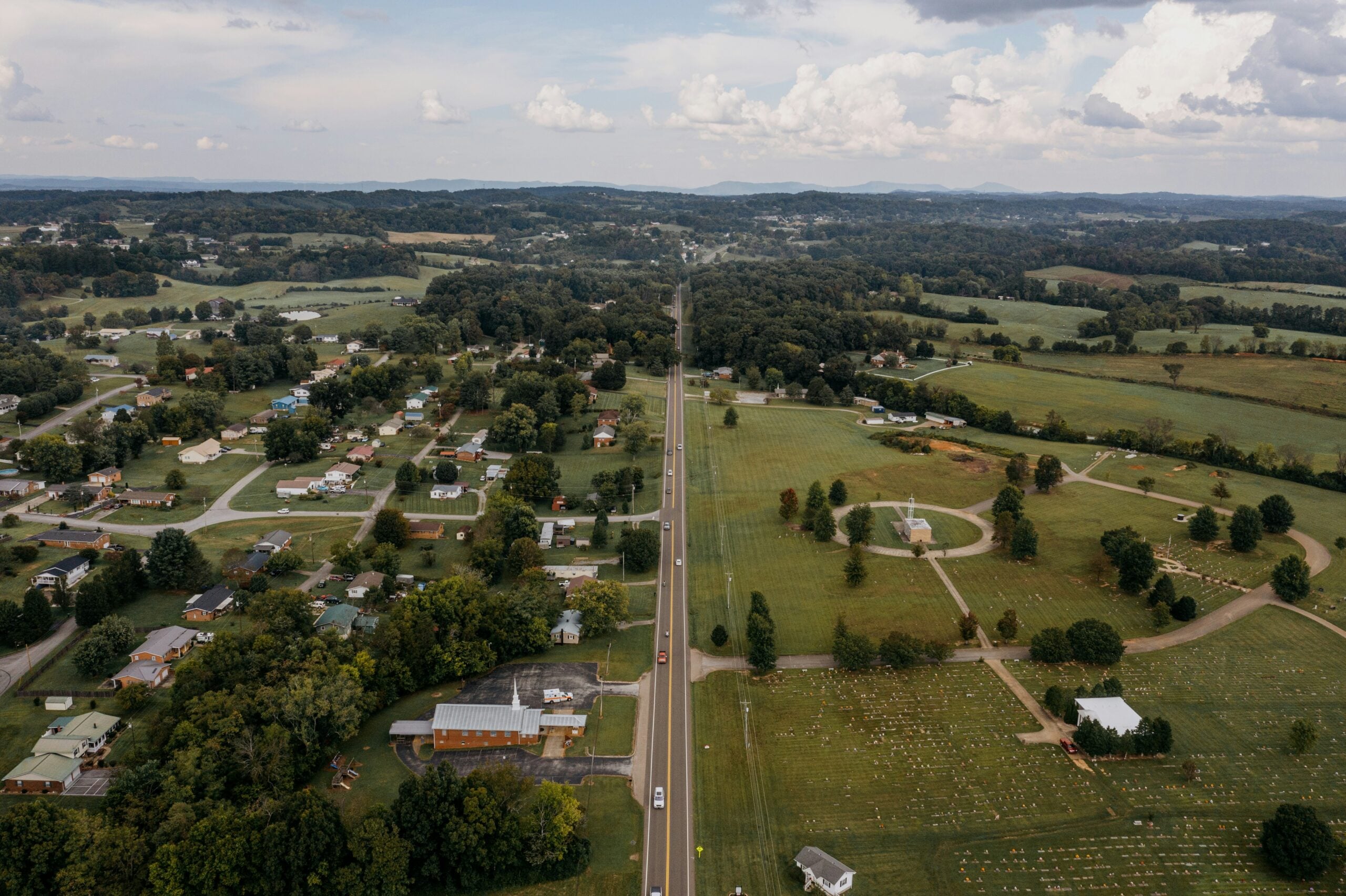 Aerial view of a straight rural road dividing a small town with houses on one side and open green fields with scattered trees and some buildings on the other under a partly cloudy sky.