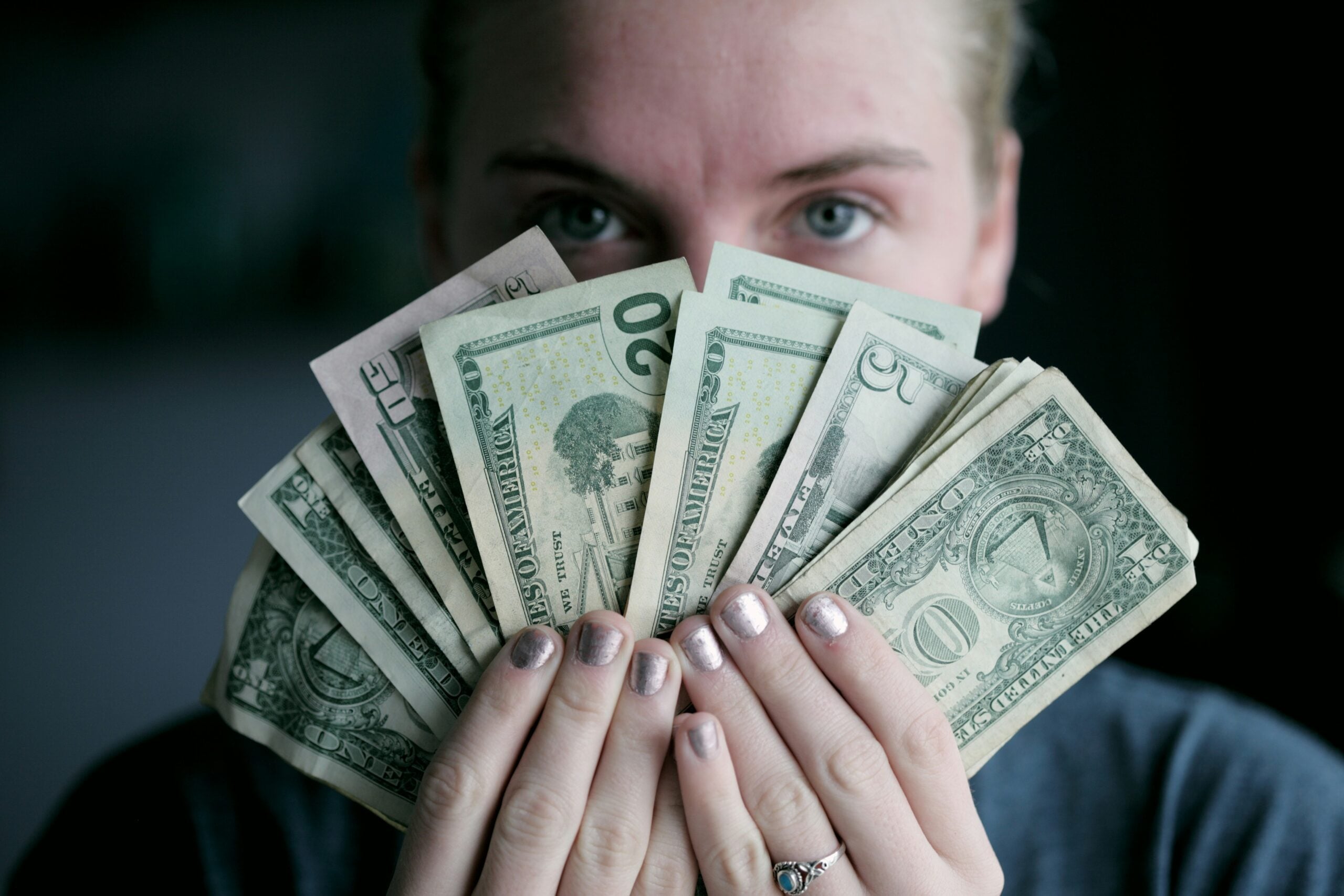 A person with light nail polish holds several U.S. dollar bills in front of their face, fanning out the money as if celebrating much-needed Cost of Living Relief and partially obscuring their features.