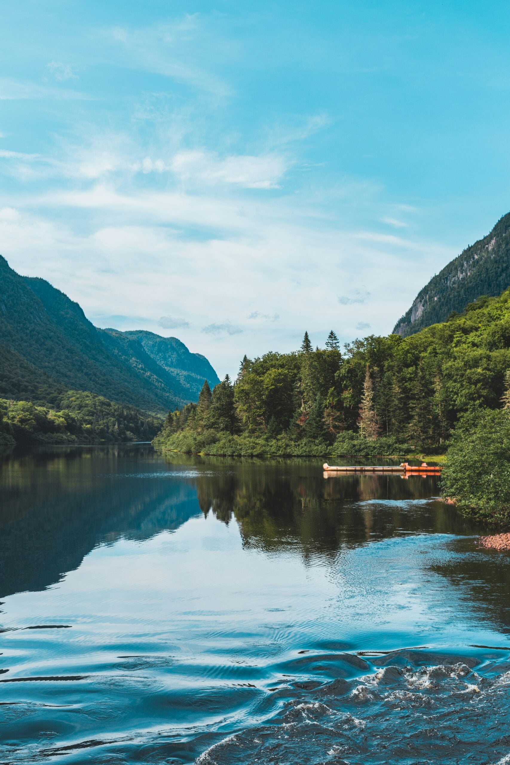 A calm river surrounded by lush green trees and mountains, with a clear blue sky above. Two orange canoes are docked at the riverbank, and the water reflects the landscape and sky.