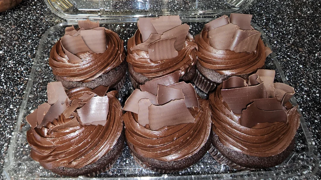 Six chocolate cupcakes sit in a clear plastic container. Each cupcake is topped with swirls of chocolate frosting and several large, curled chocolate shavings. The container is on a speckled black countertop.