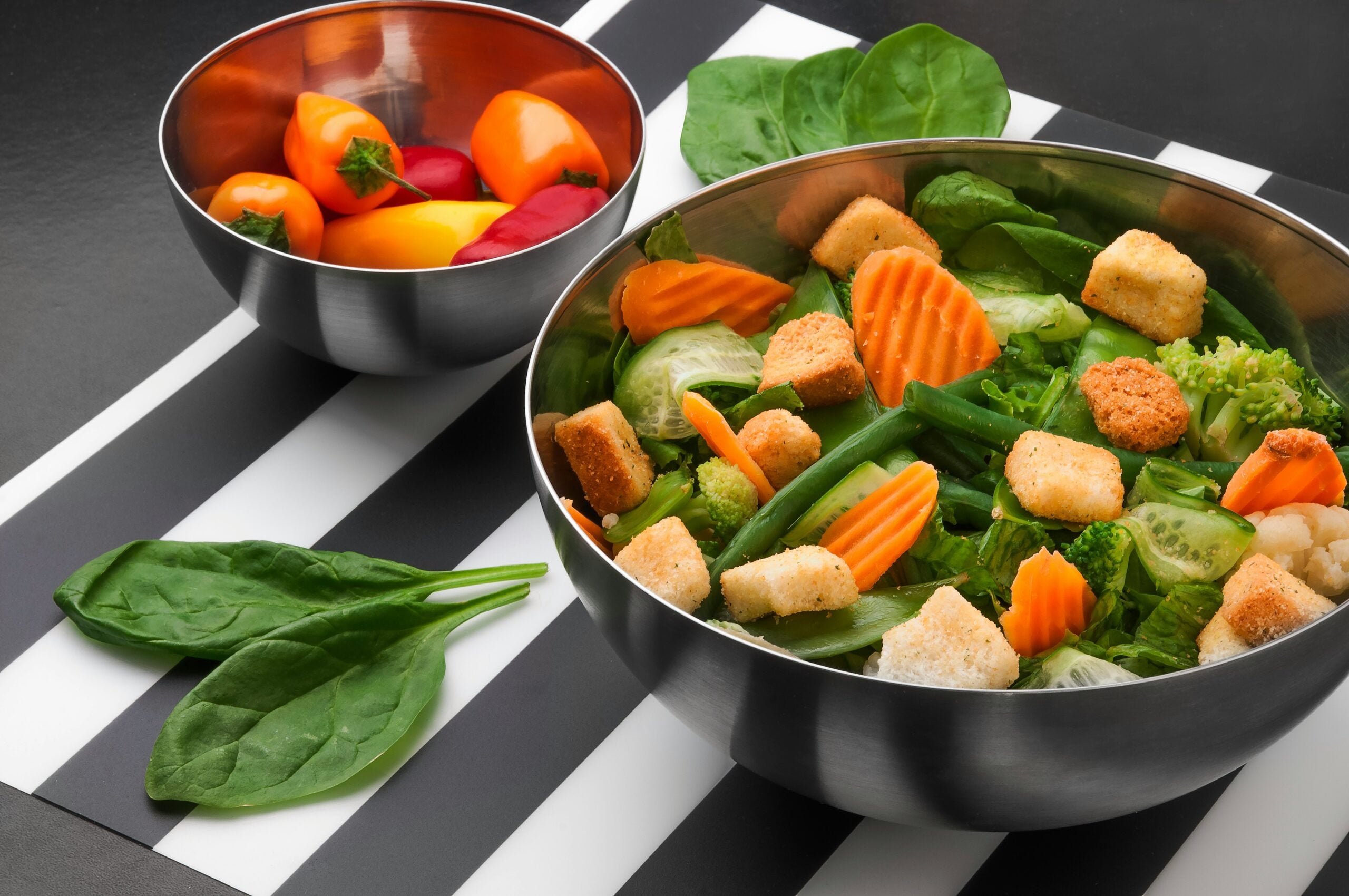 A stainless steel bowl filled with a fresh salad of greens, cucumbers, carrots, croutons, and snap peas sits beside another bowl of mini sweet peppers on a black-and-white striped surface with spinach leaves nearby.