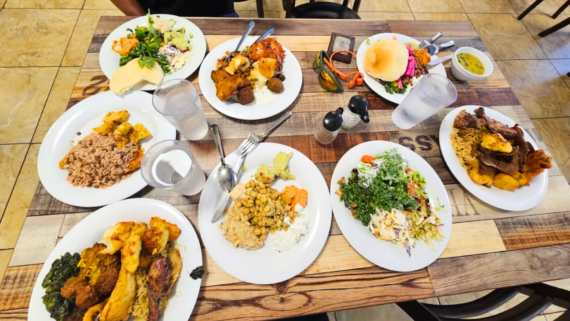 A wooden table set with plates of diverse dishes&mdash;rice, vegetables, salad, meats, a sandwich, water glasses, and utensils&mdash;suggests an inviting All You Can Eat buffet-style dining experience.