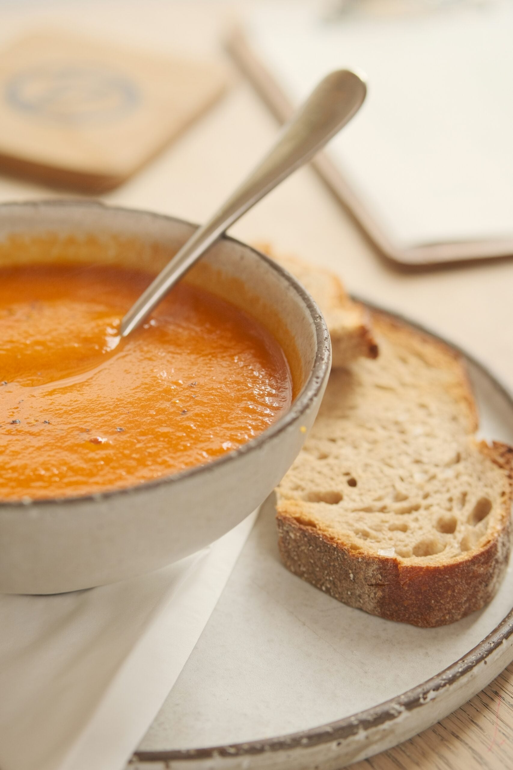 A bowl of creamy tomato soup with a spoon, served on a plate with two slices of rustic bread on the side.