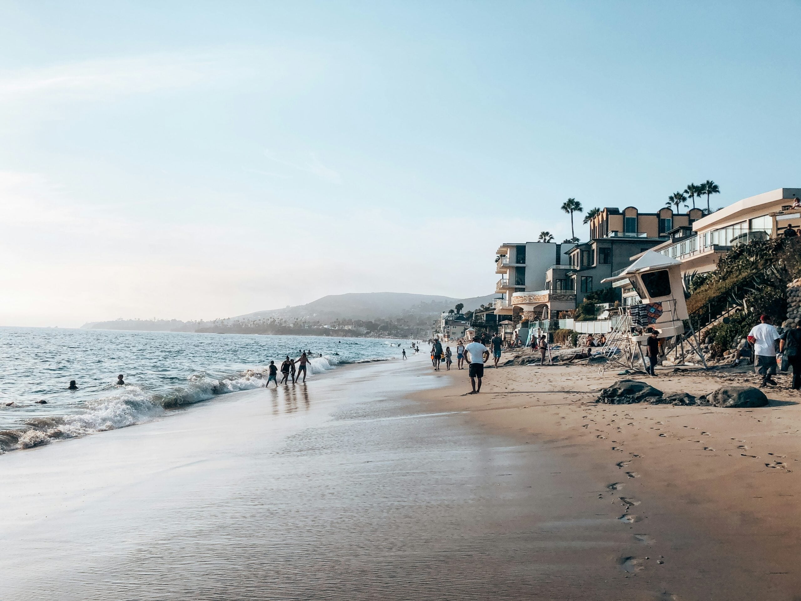 People walk along a sandy beach near ocean waves, with modern beachfront houses and palm trees lining the shore under a clear blue sky. Hills are visible in the background.