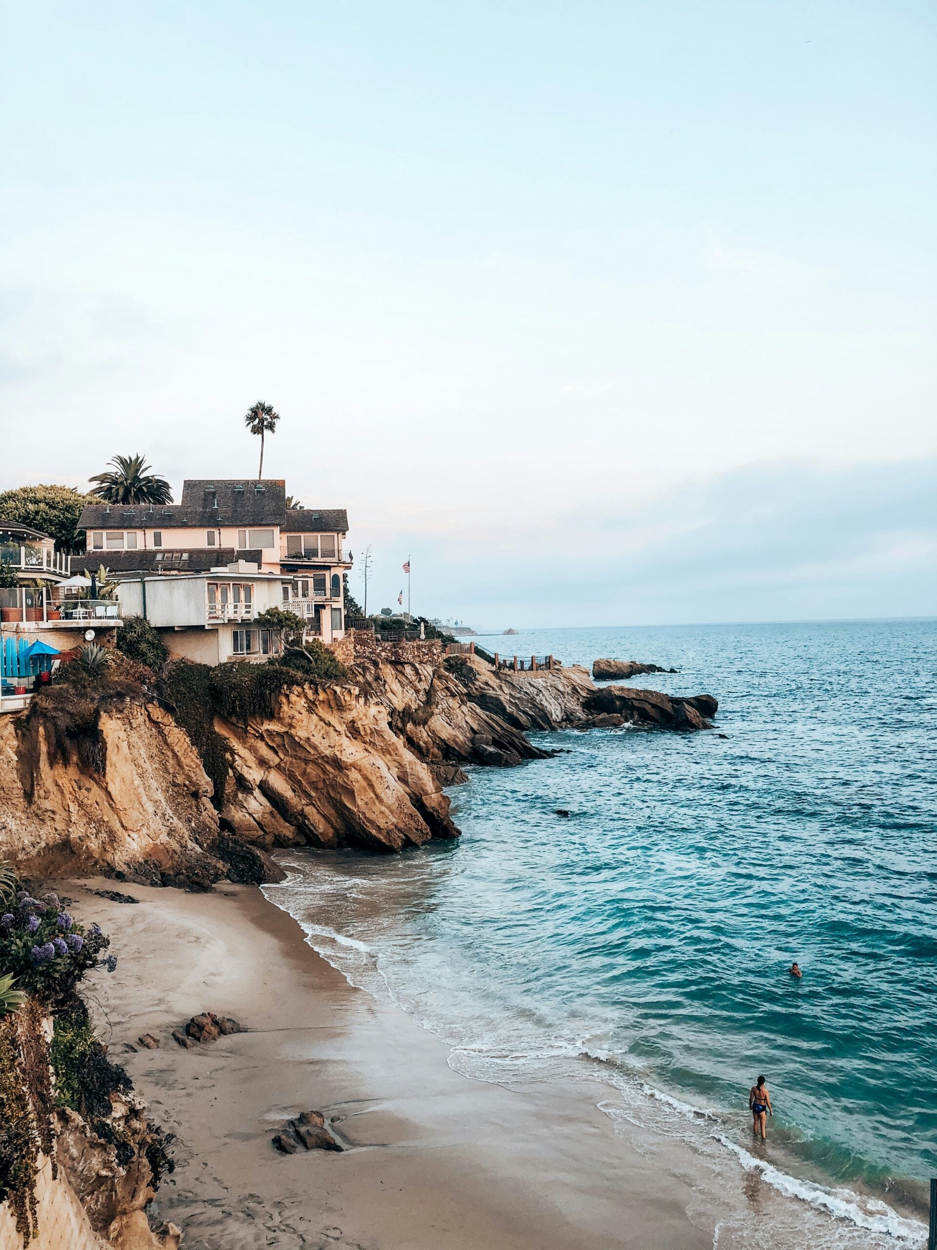 Houses sit atop rocky cliffs overlooking a sandy beach and blue ocean, with gentle waves. One person swims in the water near shore under a clear, lightly clouded sky.